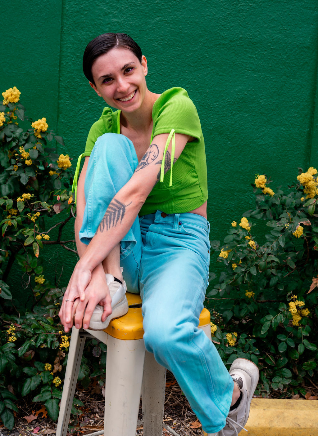 A smiling woman is sitting on a yellow stool mid laugh. She is wearing a neon green shirt and bright blue pants. There is a forest green wall behind the woman as well as yellow flower bushes.